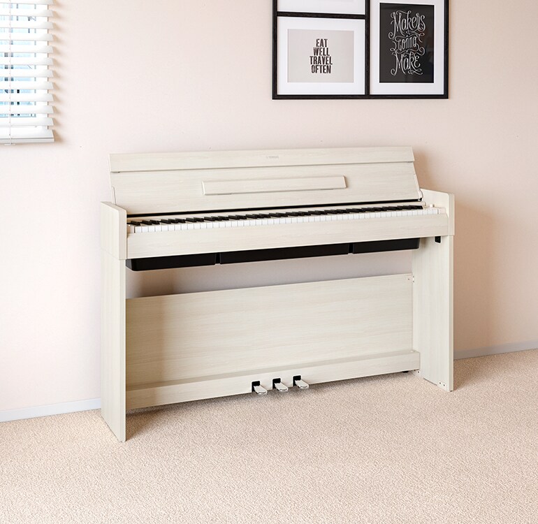 A living room featuring a white birch ARIUS piano placed next to wall shelves on which lamps and a diffuser are placed, hamoniously  integrated with the interior.